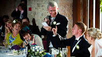 Father of the bride prepares to deliver his speech, but is interrupted by the groom holding up a finger. However, everyone is smiling.